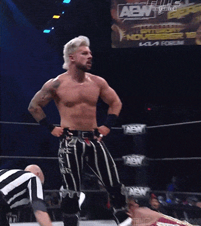 a man is standing in a wrestling ring with his hands on his hips while a referee watches .