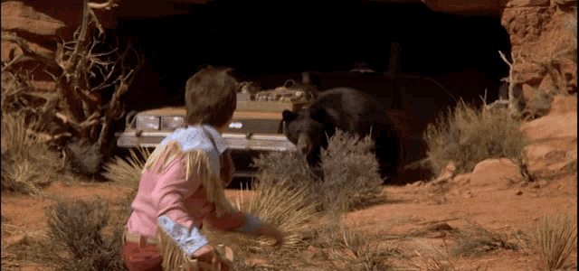 a man in a pink shirt is kneeling next to a bear in the desert