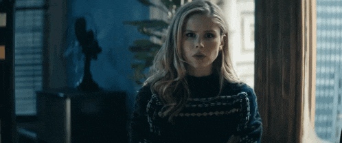 a woman in a black sweater stands in front of a window with her arms crossed