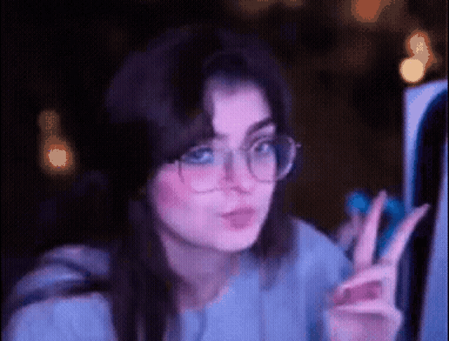 a woman wearing glasses is giving a peace sign while looking at a computer screen .