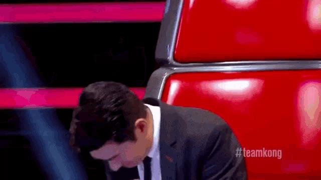 a man in a suit and tie is sitting in a red chair with #teamkong written on the bottom