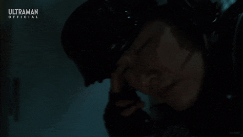 a man in a helmet is talking on a cell phone with the ultraman official logo in the corner