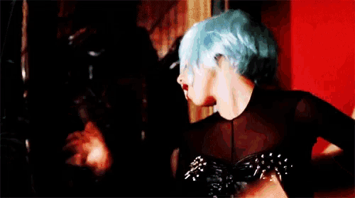 a woman with blue hair is wearing a black dress and standing in front of a red wall .