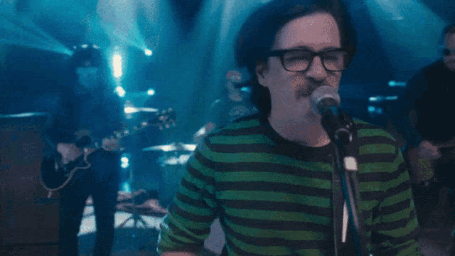 a man singing into a microphone wearing glasses and a green and black striped shirt