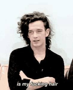 a man with long curly hair is sitting with his arms crossed and says is my fucking hair