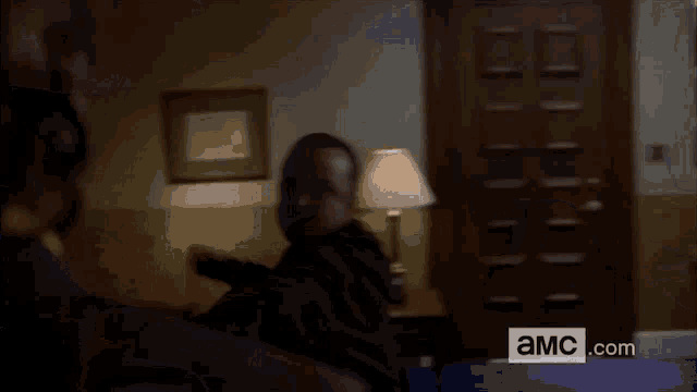 a man is sitting in a room with a lamp and a sign that says ' a.m.c.com '