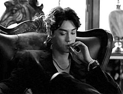 a black and white photo of a man smoking a cigarette while sitting in a chair .