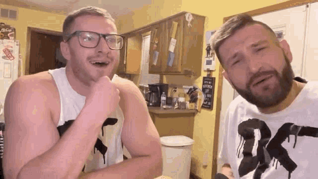 two men are standing next to each other in a kitchen . one of the men is wearing glasses and a white tank top .
