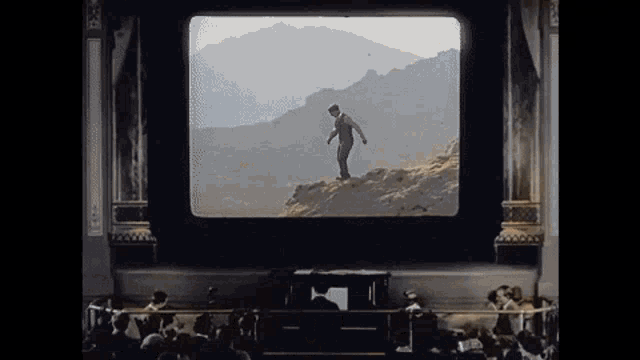 a man is standing on a cliff in front of a screen with mountains in the background