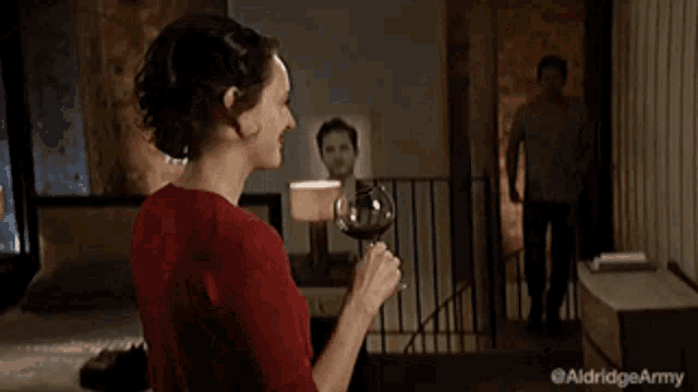a woman in a red shirt is holding a glass of wine while a man stands behind her .
