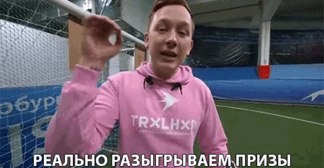 a man wearing a pink trxlhxp hoodie is standing in front of a soccer field