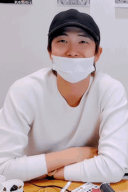 a man wearing a face mask and a hat sits at a table with his arms crossed