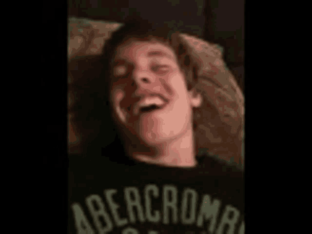 a young man wearing a black abercrombie shirt is laying on a bed laughing .