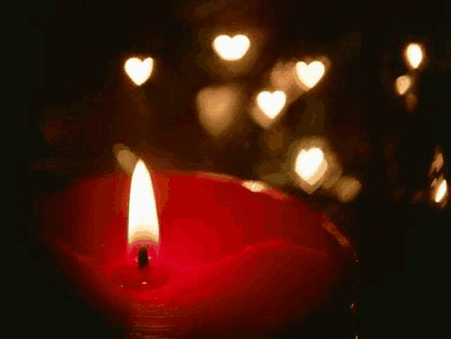 a red candle is lit up in the dark with hearts behind it .