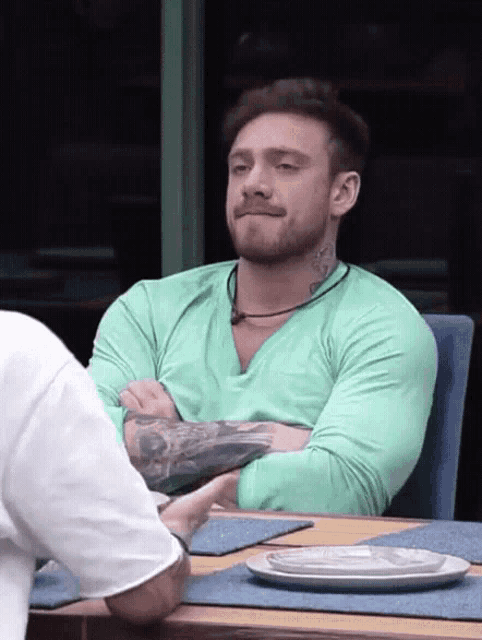 a man with a tattoo on his arm sits at a table with his arms crossed
