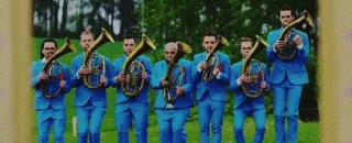 a group of men in blue suits are standing in a field playing brass instruments .