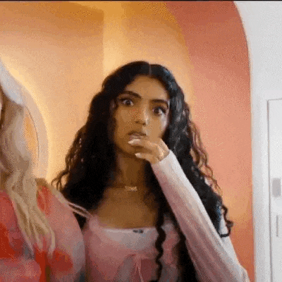 a woman with long curly hair is making a surprised face while standing next to another woman in a room .