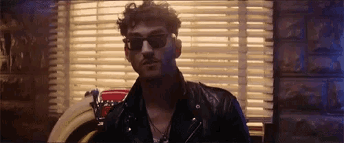 a man wearing sunglasses and a leather jacket is standing in front of a window