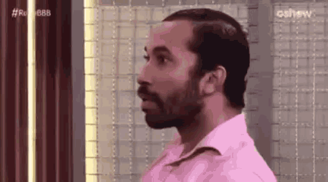 a man with a beard wearing a pink shirt is standing in front of a wall .