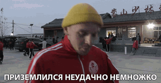 a man wearing a yellow beanie and a red jacket is standing in front of a building with chinese characters on it
