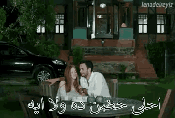 a man and woman are sitting at a table in front of a house with a car parked in the background .