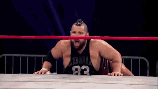 a wrestler in a black tank top with the number 83 on it is sitting in a wrestling ring .