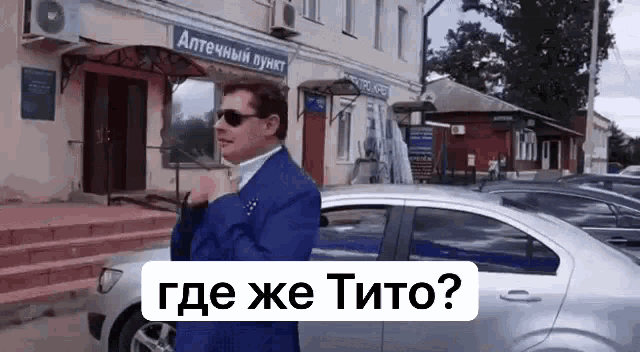 a man in a blue suit and sunglasses is standing next to a silver car .