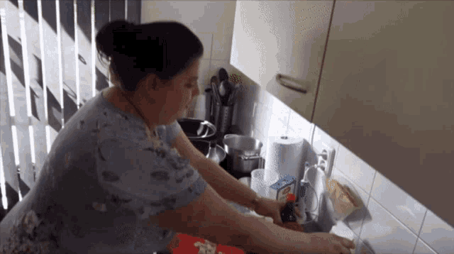 a woman in a blue shirt is washing dishes in a kitchen with a carton of aquafina on the counter