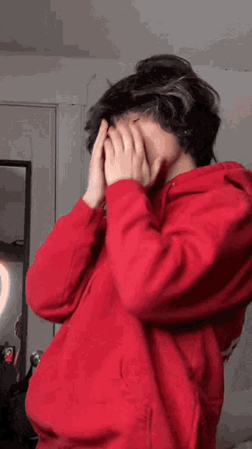 a young man wearing a red hoodie is covering his face with his hands .