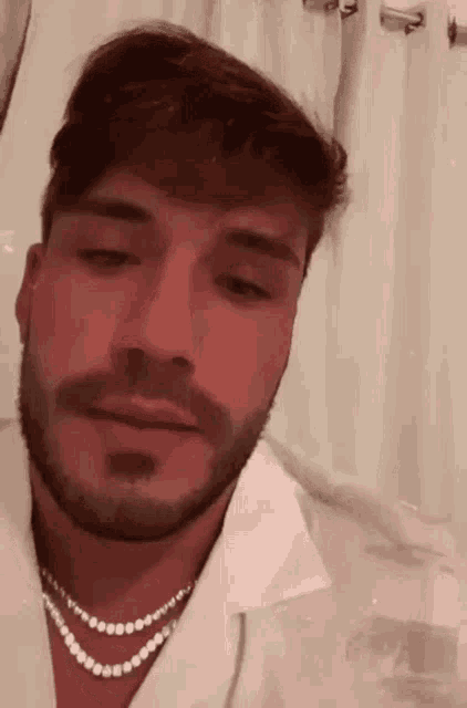 a man with a beard and a pearl necklace is wearing a white shirt and looking at the camera .