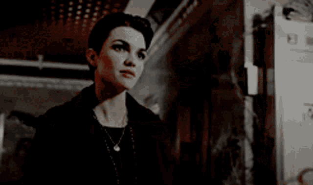 a woman wearing a black jacket and a necklace stands in a dark room .