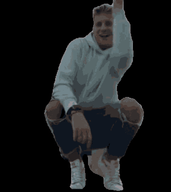 a man in a white hoodie and ripped jeans is squatting down with his arms in the air