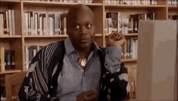 a man is sitting in front of a computer in a library and making a funny face .