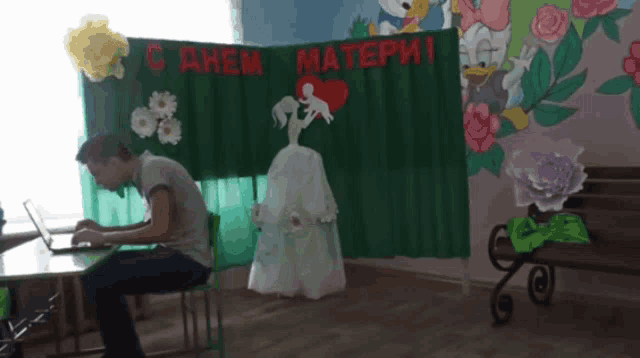 a man sits at a desk in front of a green curtain that says " c днем матери " on it