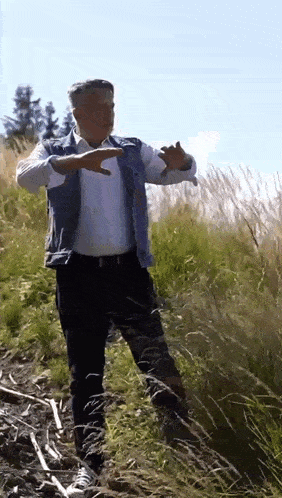 a man in a denim vest is standing in a field of tall grass making a funny face .