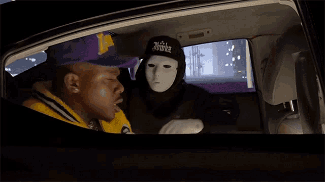 a man wearing a purple lakers hat sits in a car next to another man wearing a mask