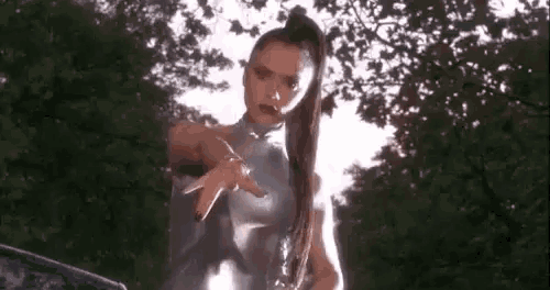 a woman in a silver dress is standing in front of a tree and pointing at the camera .