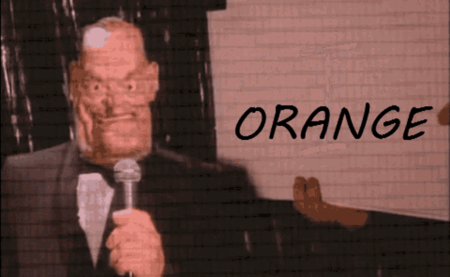 a man in a tuxedo holds a microphone in front of a wall that says orange