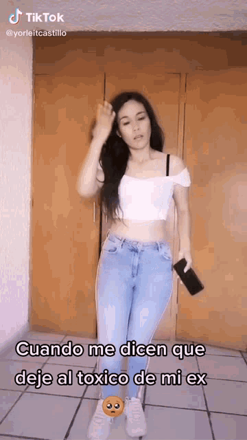 a woman in a white off the shoulder top and blue jeans is holding a cell phone and dancing .
