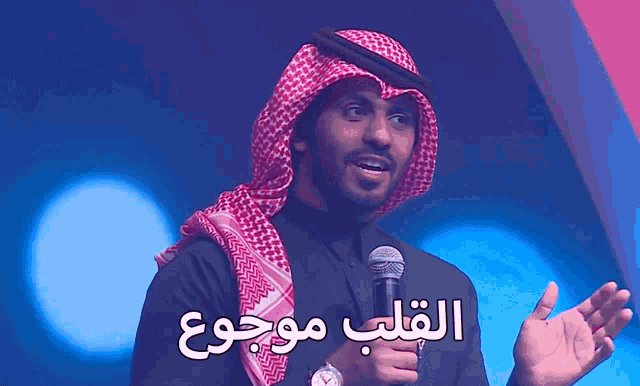a man in a keffiyeh is holding a microphone with arabic writing behind him