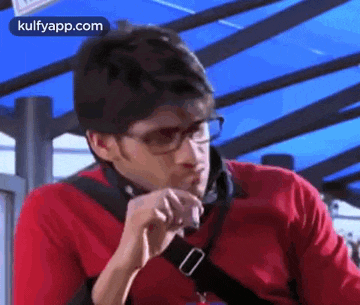 a man wearing glasses and a red shirt is smoking a cigarette while sitting in front of a blue canopy .