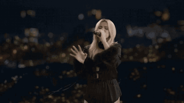 a woman is singing into a microphone in front of a city at night