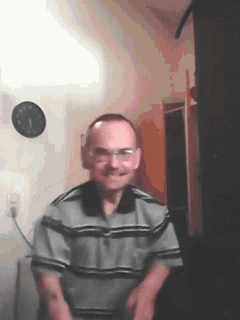 a man wearing glasses and a striped shirt is smiling in front of a clock on the wall