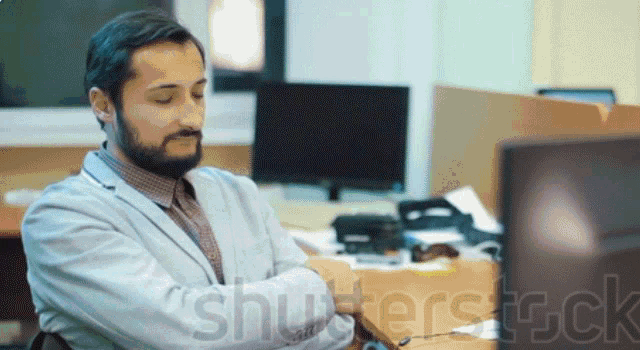 a man sits at a desk with his arms crossed in front of a computer screen with the watermark shutterstock visible