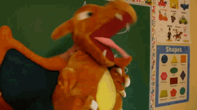 a stuffed animal is standing in front of a chalkboard with a poster that says shapes on it