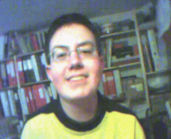 a young man wearing glasses and a yellow sweater smiles in front of a bookshelf