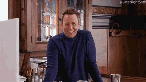 a man in a blue sweater is smiling in front of a stained glass cabinet that says late night seth
