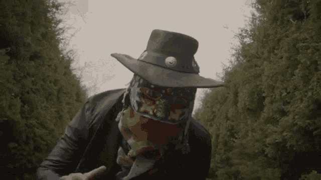 a man wearing a cowboy hat and a scarf around his face is standing in a forest .