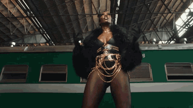 a woman in a black fur coat and gold chains stands in front of a green train