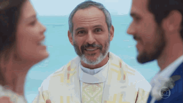 a priest smiles while a bride and groom look on during a wedding ceremony